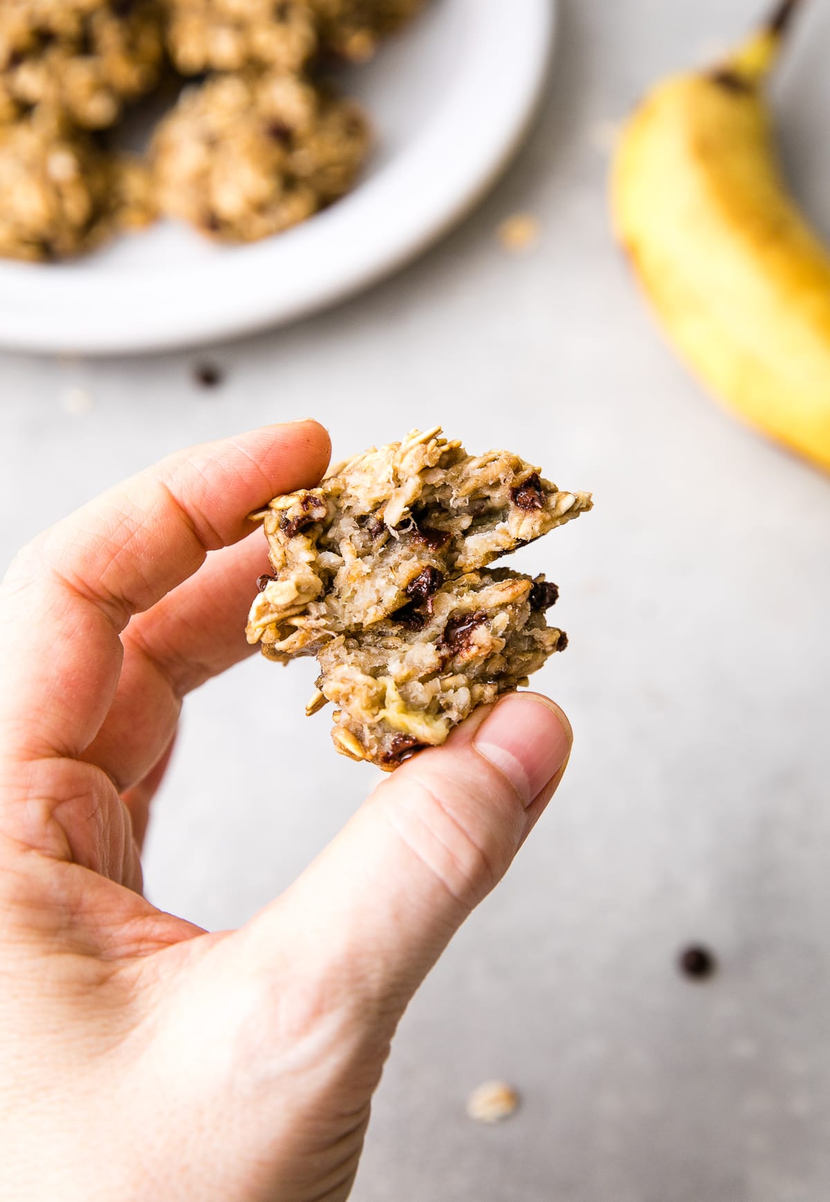 top down view of hand holding 3 ingredient banana oatmeal cookies split in half with items in background.